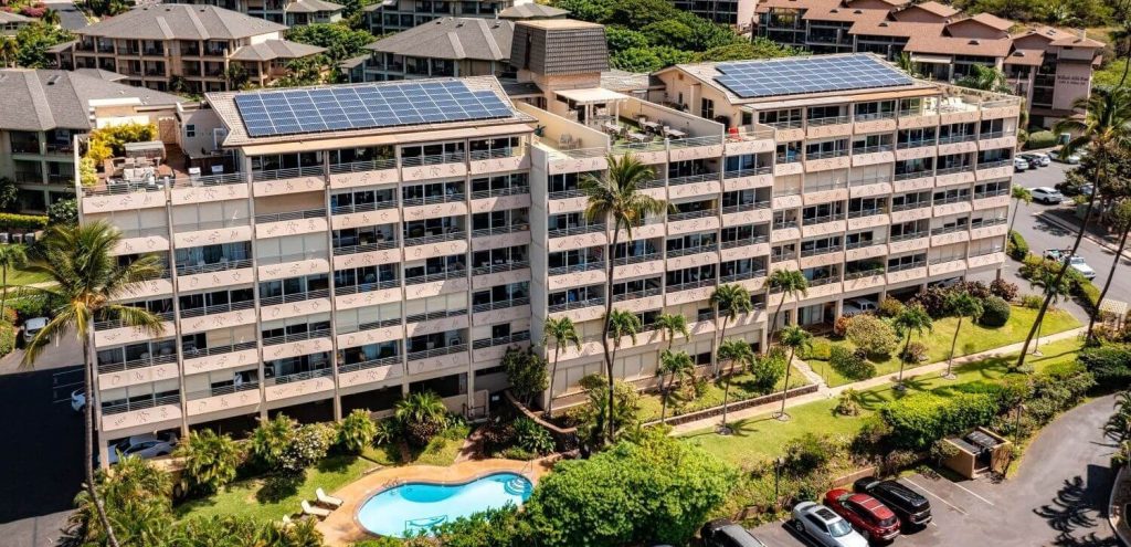 Aerial view of Kamaole Beach Royale in South Kihei, Maui, with rooftop solar panels, gated pool, lush landscaping, and six-story ocean-facing units.